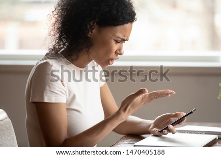 Head shot side view angry african american young woman sitting at table, holding cellphone, irritated by broken device, malware, poor internet connection, low battery, received spam message, bad news. Royalty-Free Stock Photo #1470405188