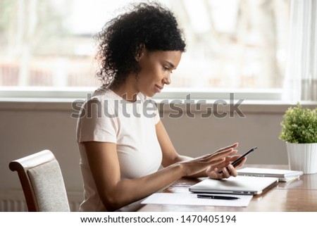 Distracted from paper work or studying young calm african ethnic millennial woman sitting at table, holding smartphone, watching editing photos for social network, typing message, using mobile apps.