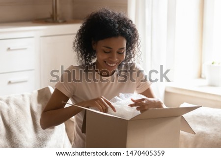 Smiling happy african athnicity young woman sitting on couch in living room, unpacking carton box parcel online order from international internet store, satisfied with fast safe air delivery service. Royalty-Free Stock Photo #1470405359