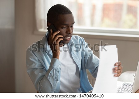 Focused young african american man holding paper documents, calling to financial advisor or bank call center support, consulting about investment, negotiating clauses of contract, trouble solving.