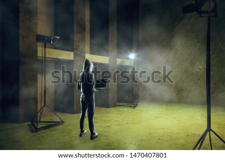 Hacker with laptop standing in green smoky concrete interior with lighting equipment.