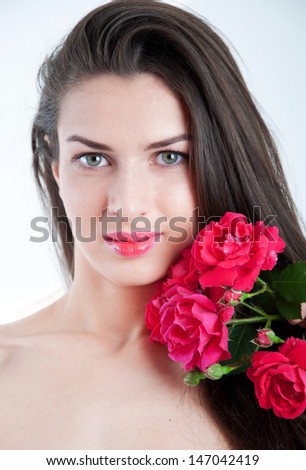Young pretty girl model with healthy long straight brunette hair, natural makeup tender glossy lips, gray eyes, holding twig with red roses near face, looking at camera, smiling. Light gray background
