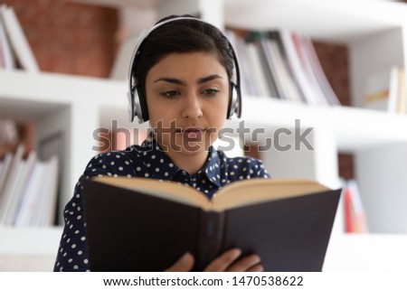 Close up head shot focused indian female student reading educational textbook, listening to favorite music in modern wireless headphones, preparing for evaluation examination in college.