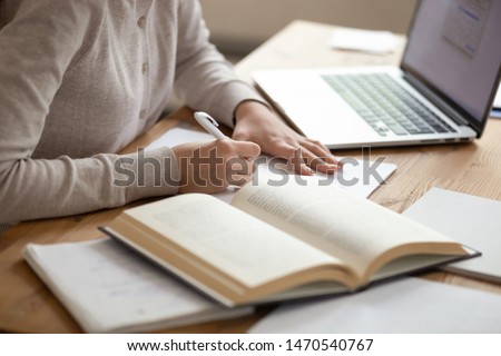 Close up female student sitting at desk, writing down notes from opened textbook and laptop. Young girl preparing for university session, exams, doing homework, paperwork, writing coursework or essay. Royalty-Free Stock Photo #1470540767