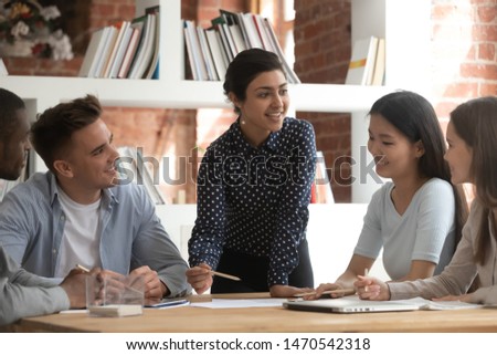 Cheerful indian female team leader involved in studying process, explaining new material to mixed race friends. Group of smiling diverse students discussing together school project ideas in classroom. Royalty-Free Stock Photo #1470542318