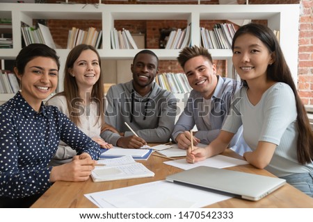 Happy mixed race students gathered in classroom, looking at camera. Smiling group of diverse young people holding video call lecture with abroad online course professor, ready to write down notes. Royalty-Free Stock Photo #1470542333