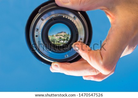 
Photographer's hand, holding photographic lens with a blue background, the focus is on the lens that reflects a medieval castle