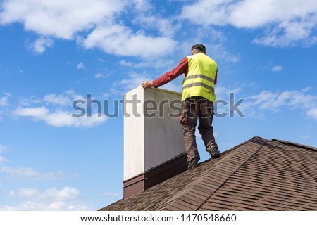 Professional workman standing roof top and measuring chimney of new house under construction against blue background Royalty-Free Stock Photo #1470548660