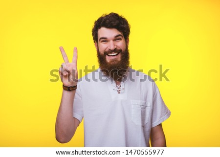 Cheerful young bearded man is showing peace sign on a yellow background.