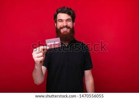 Joyful young bearded man is showing a credit card on a red background.