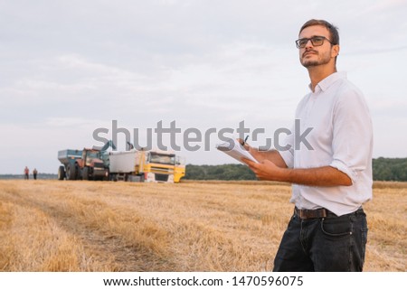 Young attractive farmer with laptop standing in wheat field with combine harvester in background