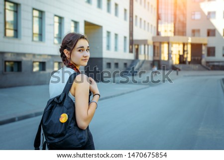 Rear view beautiful brunette with a black backpack behind going to College. A young pretty student looks back at the camera. A beautiful girl going to school. Concept of the Day of knowledge.