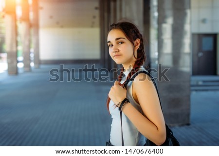 Young beautiful student with backpack on her shoulder goes to school, close-up. Schoolgirl with two braids standing in front of college, blurred background. September 1, the beginning of school year.