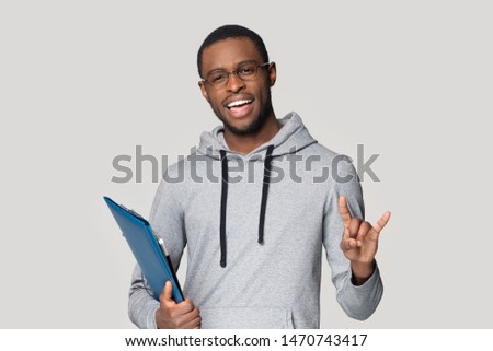 Joyful happy african american millennial male student or young professional in glasses standing with documents in folder, posing for photo, having fun, joking, isolated on grey studio background.