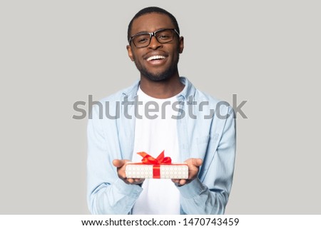Cheerful smiling african american millennial guy in eyewear holding wrapped present box head shot portrait. Happy black young man congratulating, giving birthday gift, isolated on grey background.