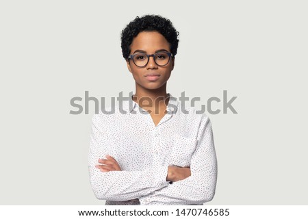 Serious concentrated young african american girl in eyeglasses standing with folded hands, headshot studio portrait. Focused black female professional looking at camera, isolated on grey background. Royalty-Free Stock Photo #1470746585