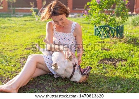 Laughing positive young woman in dress plays with her beloved restless dog sitting in the yard of a country house on a sunny summer day. Family weekend concept.