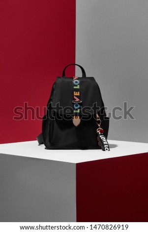 Photo of black leather backpack with strap with inscription "love" on a flap with cute teddy keychain and ribbon with inscription "love". The photo is taken on red and grey background. 
