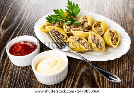 Bowls with mayonnaise and ketchup, stuffed pasta conchiglie, parsley, fork in white oval dish on dark wooden table