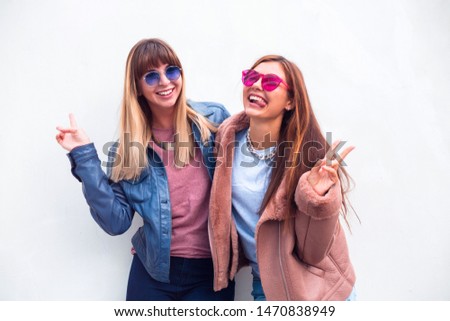 Two happy pretty pretty women in blue and pink leather jackets posing together and showing peace gestures while looking at the camera over white background outdoor.Picture of two playful girls 