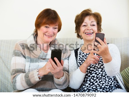 Smiling elderly friends sitting with mobile phones in living room. Focus on right woman Royalty-Free Stock Photo #1470879092