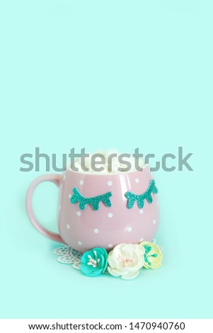 Pink mug with white polka dots with blue closed eyes with coffee and marshmallows and white, yellow and blue flowers nearby. Shiny eyelashes. Blue background.

