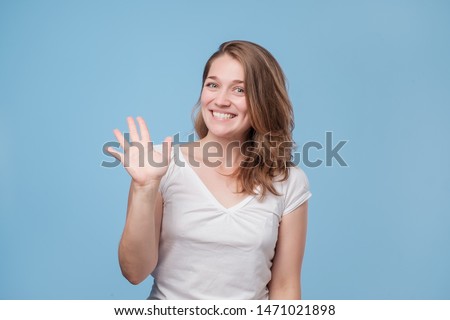 Attractive friendly looking young woman smiling happily, saying Hello, Hi or Bye, waving hand. Showing five fingers. Studio shot Royalty-Free Stock Photo #1471021898