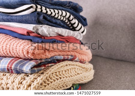 Bunch of clothing, knitted warm pastel color sweaters, different shirts and pants folded in stack on grey textile couch. Fall winter season apparel stacked on sofa. Close up, copy space, background.