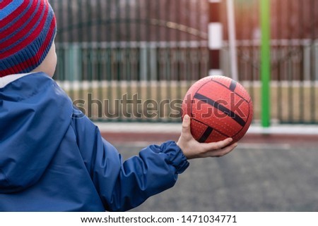 Little Boy, kid child in a blue jacket and hat holds a ball in one hand, standing on the field with a goal. Back view. Copyspace. Concept of school sports, training, outdoor activities, ball games.