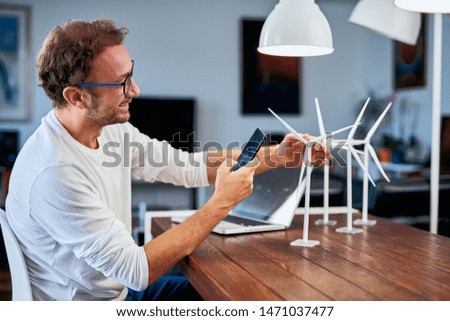 Handsome Caucasian ecology engineer sitting at home and taking photo of windmill model . On desk are windmill models. Sustainable development concept.