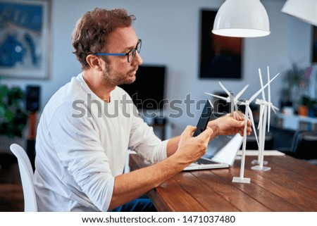 Handsome Caucasian ecology engineer sitting at home and taking photo of windmill model . On desk are windmill models. Sustainable development concept.
