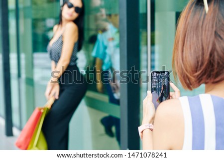 Rear view of young woman holding mobile phone and making a photo of her friend while she posing near the shop window outdoors