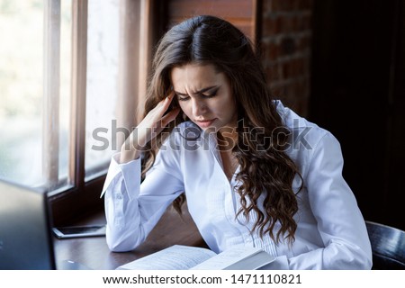 Young woman brunette with curly hair working with laptop and documents. Beautiful girl reading book near window. 
