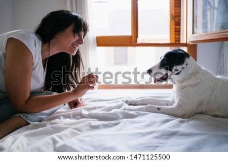 young caucasian woman on bed with her cute puppy dog playing and giving him treats. Love for animals concept. Lifestyle indoors