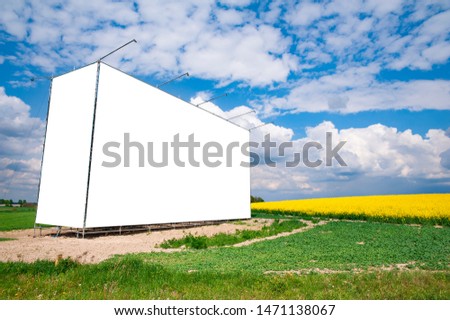 Blank white banner/billboard on the meadow near blossoming rapeseed field. Sunny day with blue sky and some white clouds.