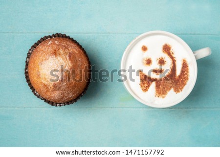 White cup of coffee with panda pattern on the foam and a cupcake on a blue wooden background, top view, copy space.