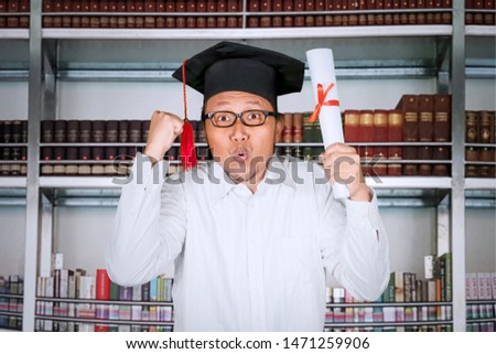 Picture of a happy man celebrating his graduation by raising hands and diploma while standing in the library