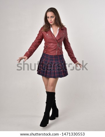 full length portrait of a brunette girl wearing a red leather jacket and plaid skirt, standing pose on a cream studio background.