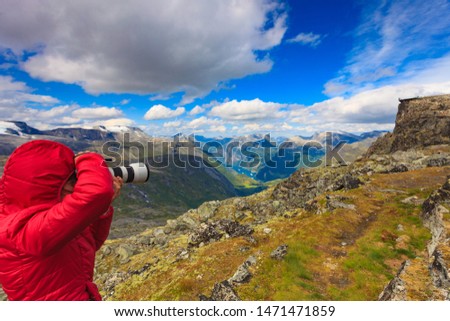 Female tourist taking photo with camera, enjoying Geirangerfjord and mountains landscape from Dalsnibba area. Geiranger Skywalk viewing platform in the distance. Norway.