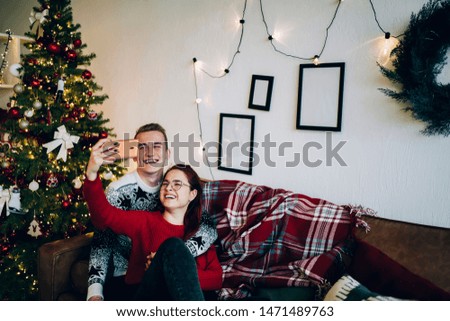 Young couple hugging and sitting on cozy sofa while smiling and taking selfie with smartphone near beautiful decorated Christmas tree during holidays