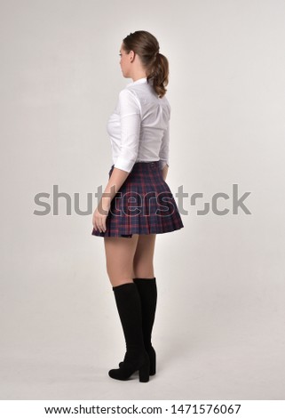 full length portrait of a brunette girl wearing a red leather jacket and plaid skirt, standing pose with back to the camera on a cream studio background