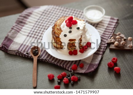 Picture of stack of pancakes with blueberries and raspberries on white plate, dark brown colour r backdrop decorated with cup of tea, spoon, pieces of chocholate and berries. Delicious breakfast.