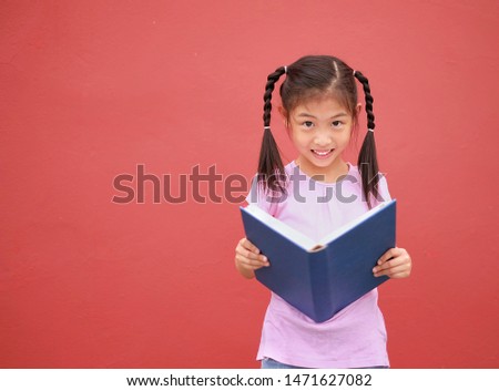 A cute Asian girl with braided hair, holding a blue text book, reading, thinking and writing with red background. Back to school