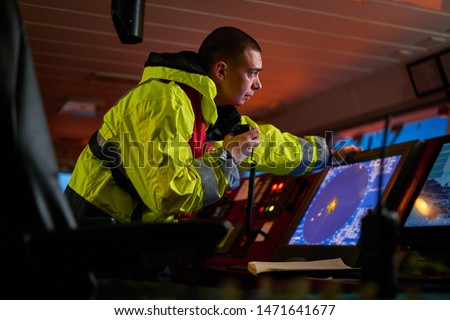 Navigator, pilot, captain part of ship crew performing daily duties with VHF radio, binoculars, logbook, standing nearby to ECDIS and radar station on board of modern ship with high quality equipment Royalty-Free Stock Photo #1471641677