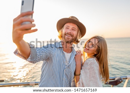 young attractive smiling happy man and woman traveling on bicycles taking selfie photo on phone camera, romantic couple by the sea on sunset, boho hipster style outfit, friends having fun together