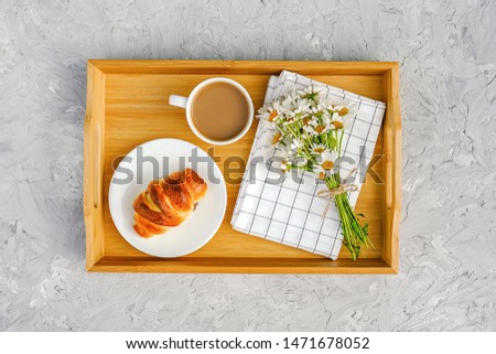 Cup of coffee with milk, freshly baked croissant, checkered napkin and camomile flowers on wooden tray on gray stone table. Concept Good morning Top view.
