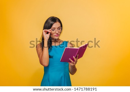 Girl with glasses on a yellow background in a blue dress reading a pink notebook