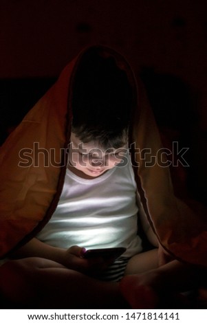 Beautiful boy hiding under the white blanket using his mobile at night time. Close up of little kid looking at smartphone secretly at night, covered with head in blanket. Technology concept. 