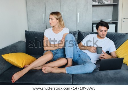 Offended blond woman sadly looking aside while brunette man with cup of coffee in hand thoughtfully using laptop. Young couple in bad mood sitting on sofa at modern home Royalty-Free Stock Photo #1471898963