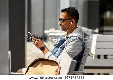 technology, travel and tourism - man with earphones, smartphone and bag sitting on city street bench and listening to music